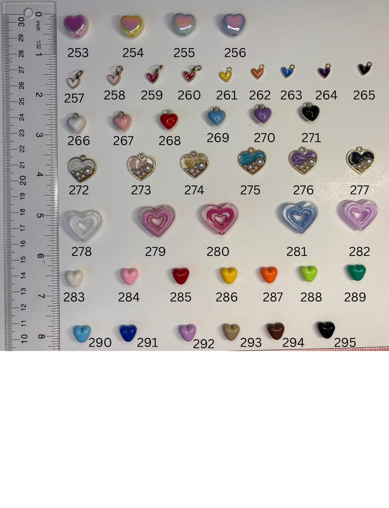May include: A collection of heart-shaped charms and beads arranged in rows, labelled with numbers 253 through 295. A clear plastic ruler is positioned on the left side, showing measurements in inches up to 8 inches for scale reference.