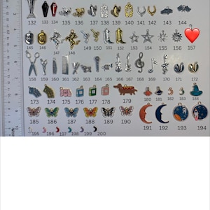 May include: A collection of metal charms numbered 132 through 200, displayed next to a ruler showing inches and centimetres. The charms include shapes like skeletons, coffins, boots, stars, musical notes, butterflies, moons, and planets in various colours.