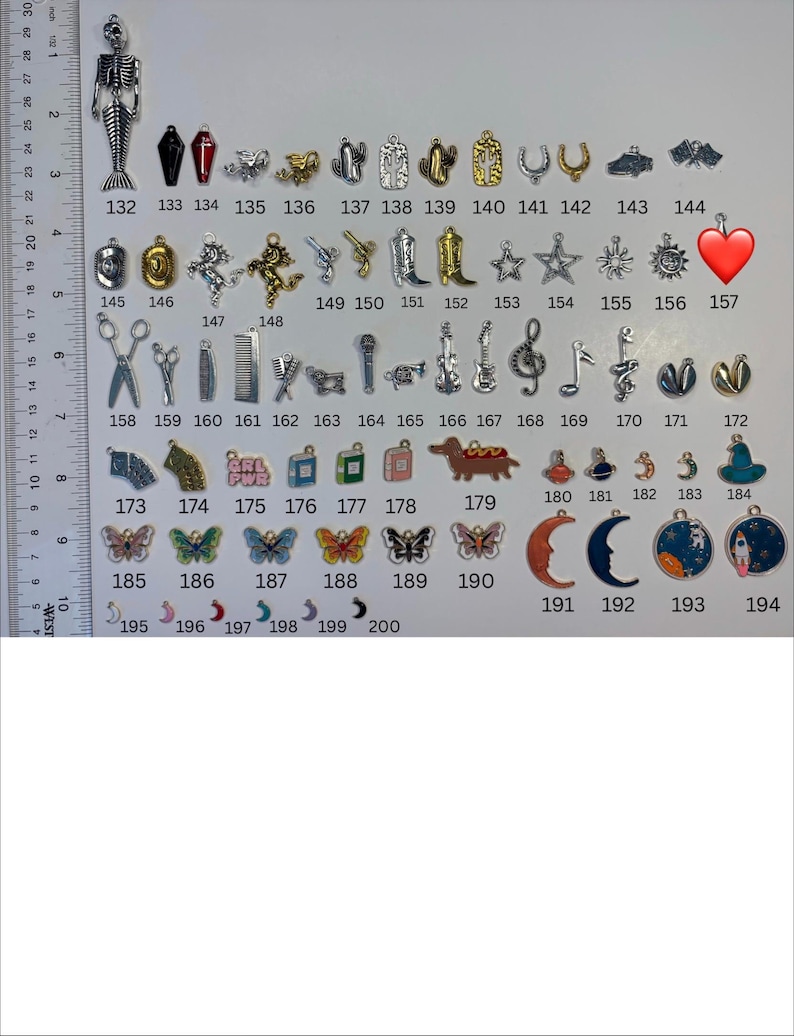 May include: A collection of metal charms numbered 132 through 200, displayed next to a ruler showing inches and centimetres. The charms include shapes like skeletons, coffins, boots, stars, musical notes, butterflies, moons, and planets in various colours.