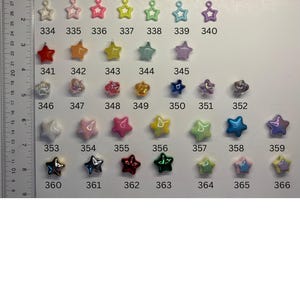 May include: A collection of small star-shaped charms in various colours and finishes, arranged in rows and numbered from 327 to 366. A ruler is positioned on the left side, showing measurements in centimetres, with the charms ranging in size.