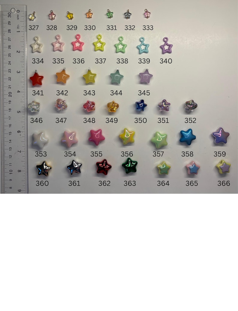May include: A collection of small star-shaped charms in various colours and finishes, arranged in rows and numbered from 327 to 366. A ruler is positioned on the left side, showing measurements in centimetres, with the charms ranging in size.