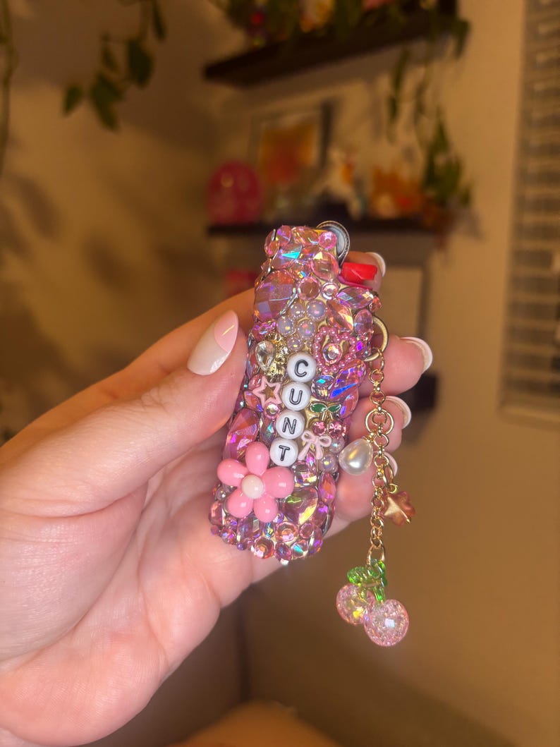 May include: A lighter case covered in pink and iridescent rhinestones, featuring a vertical row of white letter beads spelling CUNT. The case is decorated with a pink flower, a small bow, and a gold-toned chain charm with a pearl and two cherries.