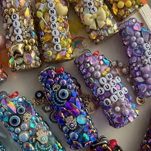 May include: A collection of lighters decorated with various colourful rhinestones, beads, and charms. Some lighters feature letter beads spelling out phrases like DIVA, SUNSHINE, and I'M JUST A GIRL, while others incorporate evil eye and hamsa hand charms.