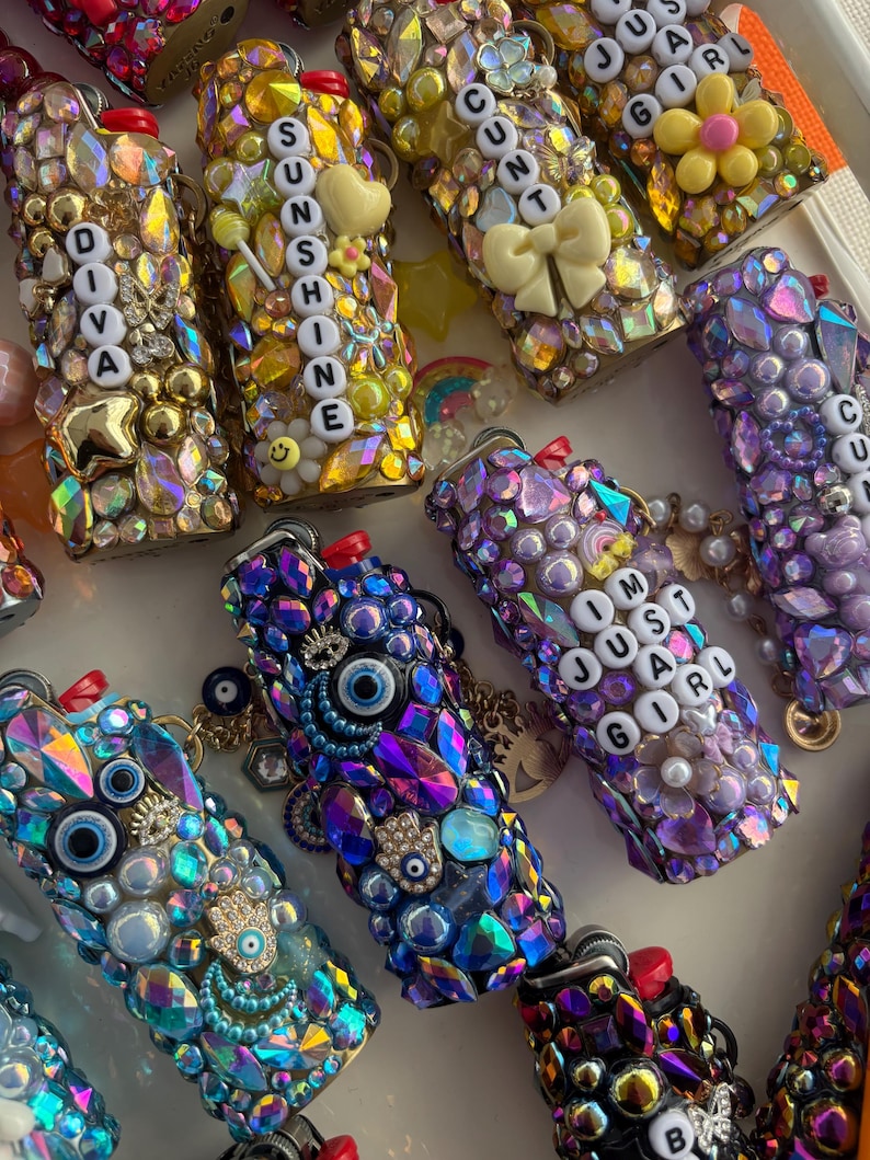 May include: A collection of lighters decorated with various colourful rhinestones, beads, and charms. Some lighters feature letter beads spelling out phrases like DIVA, SUNSHINE, and I'M JUST A GIRL, while others incorporate evil eye and hamsa hand charms.