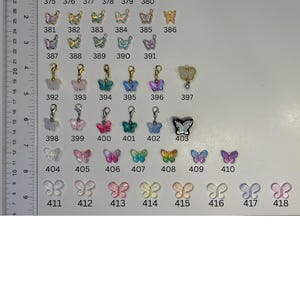 May include: A collection of small butterfly-shaped charms and pendants in various colours, finishes, and styles, arranged in rows and numbered from 367 to 418. A vertical ruler on the left side shows measurements in centimetres for scale reference.