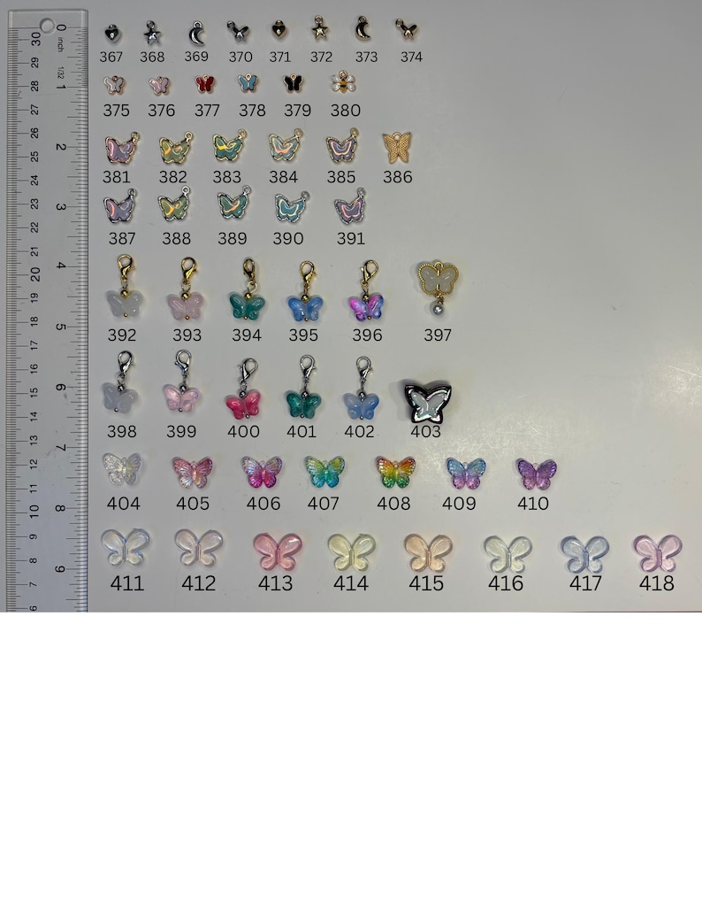 May include: A collection of small butterfly-shaped charms and pendants in various colours, finishes, and styles, arranged in rows and numbered from 367 to 418. A vertical ruler on the left side shows measurements in centimetres for scale reference.