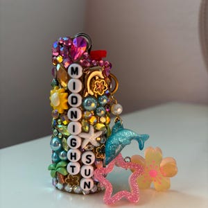 May include: A cylindrical lighter decorated with colourful rhinestones, beads, and charms. It features white letter beads spelling MIDNIGHT SUN vertically. Attached charms include a blue dolphin, a pink glittery star, and a yellow flower.