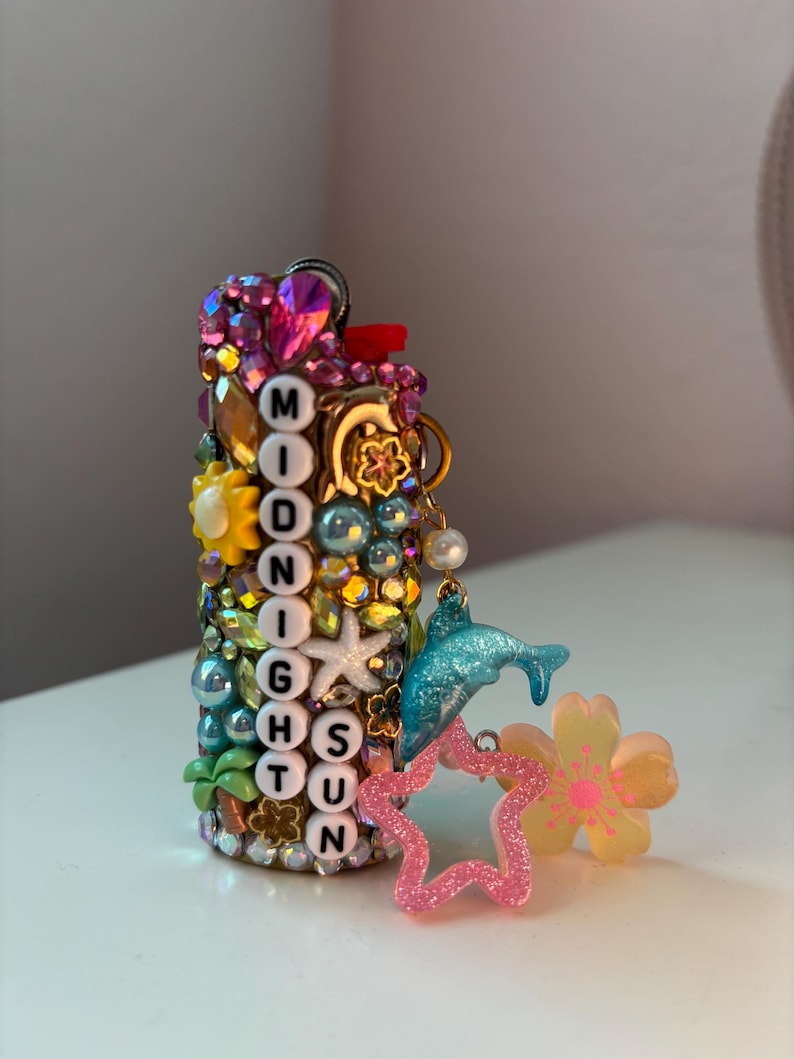 May include: A cylindrical lighter decorated with colourful rhinestones, beads, and charms. It features white letter beads spelling MIDNIGHT SUN vertically. Attached charms include a blue dolphin, a pink glittery star, and a yellow flower.