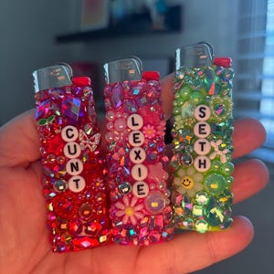 May include: Three bedazzled lighters in red, pink, and green. Each lighter is covered in rhinestones and beads, with names spelled out in white letter beads: CUNT, LEXIE, and SETH. The lighters have silver metal tops.