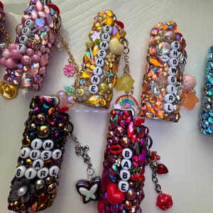 May include: A collection of five rectangular lighters covered in colourful rhinestones and charms. Each lighter features white letter beads spelling out phrases including CUNT, SUNSHINE, SHOWGIRL, IM JUST A GIRL, and BABE, with attached metal chains and charms.