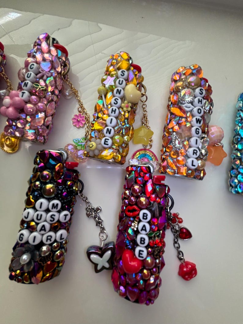 May include: A collection of five rectangular lighters covered in colourful rhinestones and charms. Each lighter features white letter beads spelling out phrases including CUNT, SUNSHINE, SHOWGIRL, IM JUST A GIRL, and BABE, with attached metal chains and charms.
