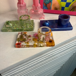 May include: Four different candle holders, each with a unique design. The first is a green holder with glitter, the second is a clear holder with dried flowers, the third is a blue holder with a round top, and the fourth is a clear holder with a pink candle.