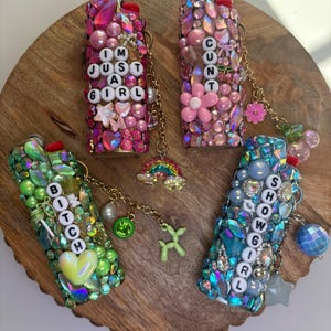 May include: Four lighters decorated with colourful rhinestones, beads, and charms. One pink lighter reads I'M JUST A GIRL, one pink reads CUNT, one green reads BITCH, and one blue reads SHOWGIRL. Each lighter features a gold chain with dangling charms.