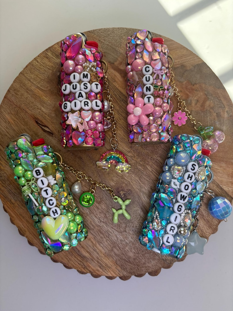 May include: Four lighters decorated with colourful rhinestones, beads, and charms. One pink lighter reads I'M JUST A GIRL, one pink reads CUNT, one green reads BITCH, and one blue reads SHOWGIRL. Each lighter features a gold chain with dangling charms.
