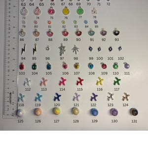 May include: A collection of numbered jewellery charms and beads arranged in rows, including smiley faces, shells, lightning bolts, balloon dogs, and spherical beads. A Westcott ruler measuring up to 30 cm is positioned vertically on the left side.