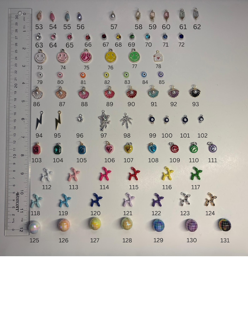 May include: A collection of numbered jewellery charms and beads arranged in rows, including smiley faces, shells, lightning bolts, balloon dogs, and spherical beads. A Westcott ruler measuring up to 30 cm is positioned vertically on the left side.