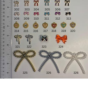 May include: A collection of metal and plastic charms in various shapes, including bows and hearts, arranged in rows with numerical labels from 296 to 326. A ruler on the left side shows measurements in cm, with the largest charms measuring 5 cm wide.