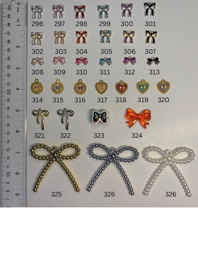 May include: A collection of metal and plastic charms in various shapes, including bows and hearts, arranged in rows with numerical labels from 296 to 326. A ruler on the left side shows measurements in cm, with the largest charms measuring 5 cm wide.