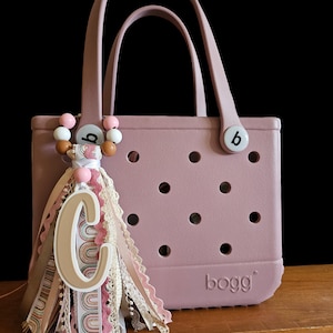 May include: A dusty rose tote bag with a decorative tassel and a letter "C" charm. The tassel features various ribbons, lace, and beads in shades of pink, cream, and brown. The bag has a handle and the brand name "bogg" is visible.