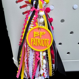 May include: A pink and yellow charm with the text "BP Patriots" hanging from a white tote bag. The charm is decorated with black, white, pink, and yellow ribbons and pom-poms.