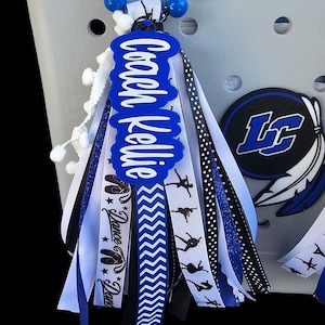 May include: A grey tote bag with a decorative charm. The charm features a blue tag with the text "Coach Kellie" and ribbons in white, black, and blue. Some ribbons have dance-themed graphics. The charm also has white and black beads.