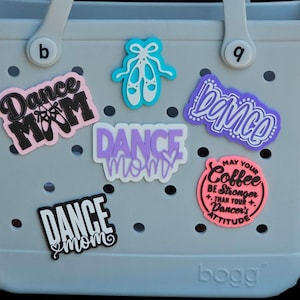 May include: A light gray tote bag with perforated holes, decorated with several colorful silicone charms. The charms feature text including Dance Mom, dance, and May your coffee be stronger than your dancer's attitude, alongside ballet shoe icons.