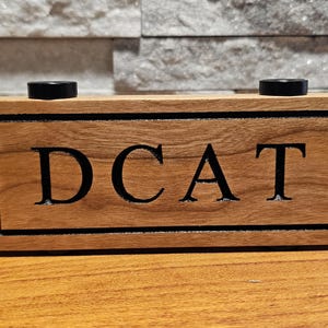May include: A wooden sign with the word "DCAT" engraved in black lettering. The sign has two black knobs on the top.
