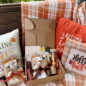May include: A cardboard gift box filled with assorted items including a glass tumbler with pumpkin prints, a small candle, a wrapped package with a brown ribbon, and stationery. The box is surrounded by orange and white plaid blankets and decorative pillows.