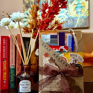 May include: A brown reed diffuser bottle with a label reading Apples & Maple Bourbon, accompanied by a tan envelope gift set labeled Cozy Mystery and Book Girl Fall Edition, featuring a brown lace ribbon, stickers, and various stationery items.
