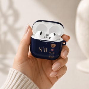 May include: A navy blue protective case for wireless earbuds featuring a graphic of a teddy bear wearing a sweater with an American flag design. The case is personalized with the gold-colored initials NB and has a white interior with two earbuds.