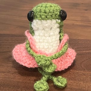 Op de afbeelding: Een handgemaakte gehaakte kikkerfiguur. De kikker is voornamelijk groen met een witte buik en een roze rok. Hij heeft zwarte kraaloogjes en groene poten. De kikker zit op een houten oppervlak.