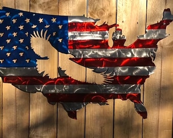 May include: Metal wall art depicting the United States map with the American flag design. The artwork features a blue section with white stars and red and white stripes, with an eagle silhouette cut out. The piece is mounted on a wooden background.