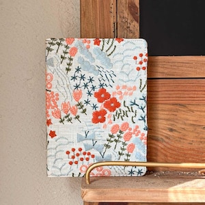 以下が含まれることがあります： 白いリネン製のノートカバーに、花柄の刺繍が施されています。刺繍には、赤、ピンク、青の花と葉が描かれています。カバーは木製の棚の上に置かれています。
