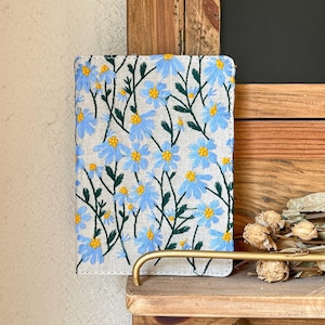 May include: A linen notebook cover with blue and yellow embroidered flowers. The cover is on a wooden shelf with a gold metal handle.