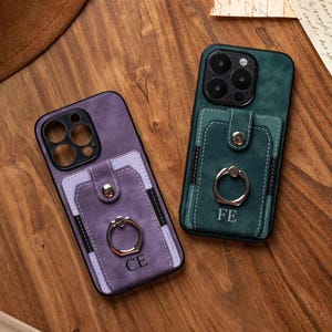 May include: Two phone cases with cardholder pockets and ring holders. One case is purple with "CE" engraved, the other is green with "FE". Both cases are on a wooden surface.