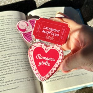 Könnte beinhalten: Ein klarer Acryl-Lesezeichen mit einem roten Herz-Design mit der Aufschrift "Romance girlie" und einem roten Ticket mit der Aufschrift "LATENIGHT BOOK CLUB". Das Lesezeichen liegt über einem offenen Buch.