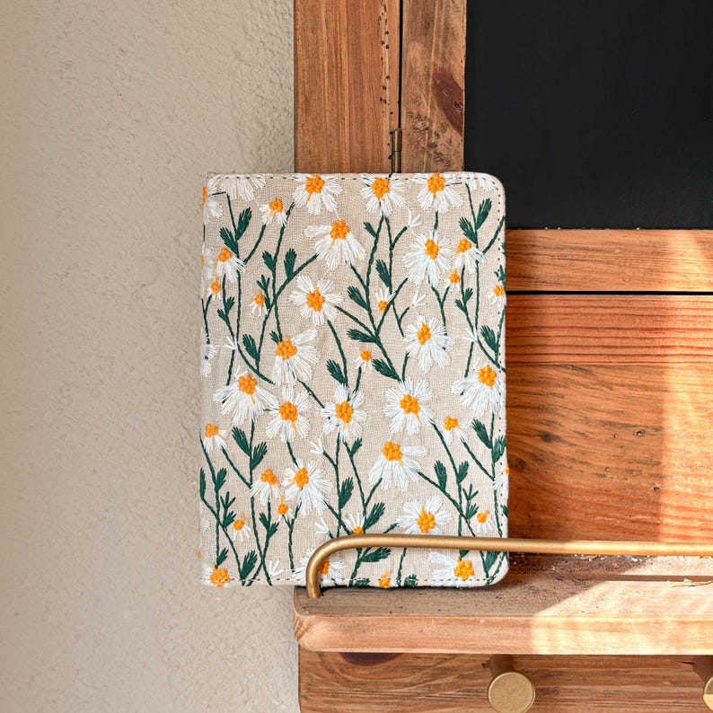 May include: A beige linen notebook cover with a white daisy floral embroidery pattern. The cover is on a wooden shelf.