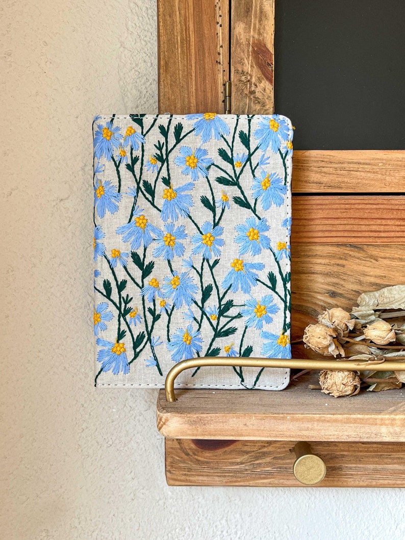 May include: A linen notebook cover with blue and yellow embroidered flowers. The cover is on a wooden shelf with a gold metal handle.