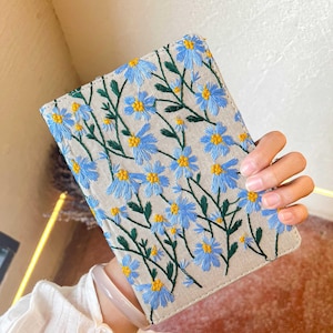 May include: A small, light beige linen notebook with blue and yellow embroidered flowers. The notebook has a simple design and is perfect for journaling or taking notes.