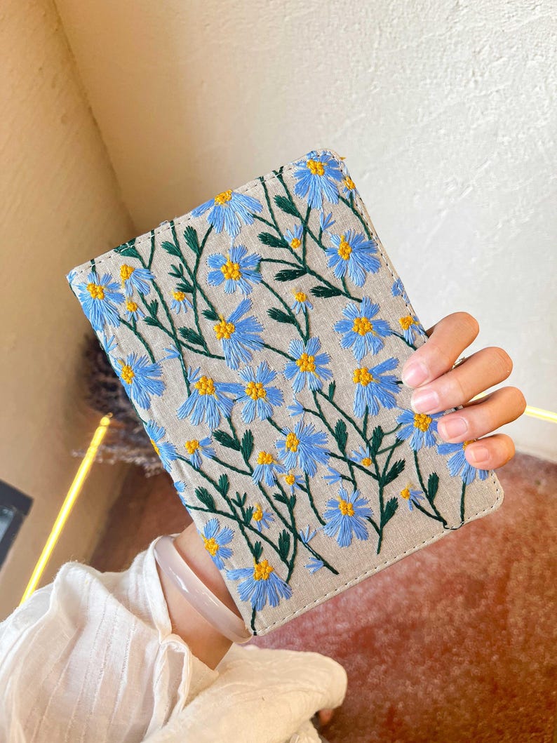 May include: A small, light beige linen notebook with blue and yellow embroidered flowers. The notebook has a simple design and is perfect for journaling or taking notes.