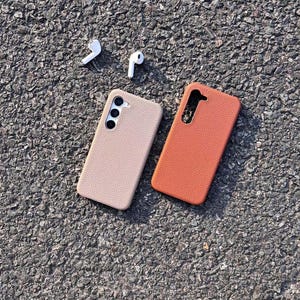Puede incluir: Dos fundas para tel&eacute;fono, una beige y otra naranja, hechas de cuero texturizado. Las fundas est&aacute;n sobre una superficie de asfalto gris. Tambi&eacute;n hay dos auriculares inal&aacute;mbricos blancos sobre la superficie.