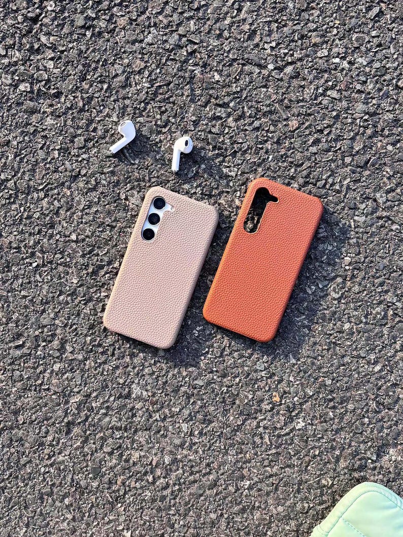 Puede incluir: Dos fundas para tel&eacute;fono, una beige y otra naranja, hechas de cuero texturizado. Las fundas est&aacute;n sobre una superficie de asfalto gris. Tambi&eacute;n hay dos auriculares inal&aacute;mbricos blancos sobre la superficie.