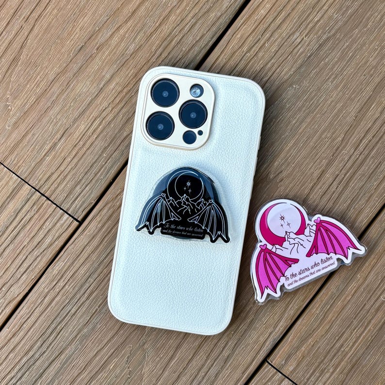 May include: A white smartphone with a black and a pink phone grip. The black grip features a bat wing design with a moon and stars, and the text "To the stars who listen and the dreams that are answered." The pink grip has the same design.