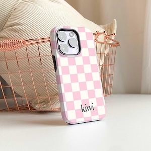 May include: A pink and white checkered phone case with the word "kiwi" in black script. The case has a camera cutout and is set against a rose gold wire basket and a white surface.