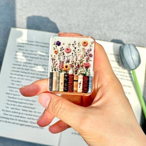 May include: A rectangular clear acrylic bookmark featuring a colorful floral design above a row of miniature books. The bookmark is held in a hand, with a book and a decorative tulip in the background.