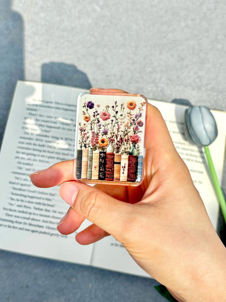 May include: A rectangular clear acrylic bookmark featuring a colorful floral design above a row of miniature books. The bookmark is held in a hand, with a book and a decorative tulip in the background.