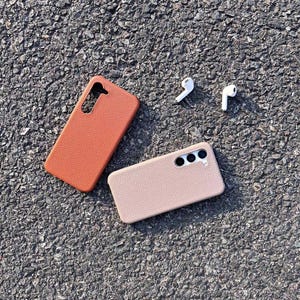 Puede incluir: Dos fundas para tel&eacute;fono, una marr&oacute;n y otra beige, hechas de cuero texturizado. Las fundas est&aacute;n sobre una superficie de asfalto gris. Tambi&eacute;n hay dos auriculares inal&aacute;mbricos blancos sobre la superficie.
