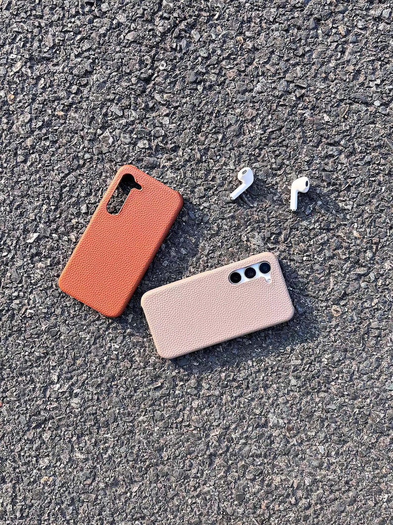 Puede incluir: Dos fundas para tel&eacute;fono, una marr&oacute;n y otra beige, hechas de cuero texturizado. Las fundas est&aacute;n sobre una superficie de asfalto gris. Tambi&eacute;n hay dos auriculares inal&aacute;mbricos blancos sobre la superficie.