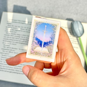 May include: A small, rectangular, white trinket box featuring a fantasy scene. The artwork depicts a sword piercing an open book, set against a backdrop of a blue sky, pink flowers, and a forest. The box is held in a hand.
