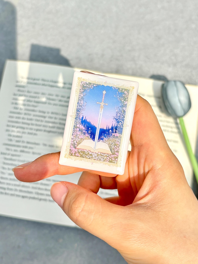 May include: A small, rectangular, white trinket box featuring a fantasy scene. The artwork depicts a sword piercing an open book, set against a backdrop of a blue sky, pink flowers, and a forest. The box is held in a hand.